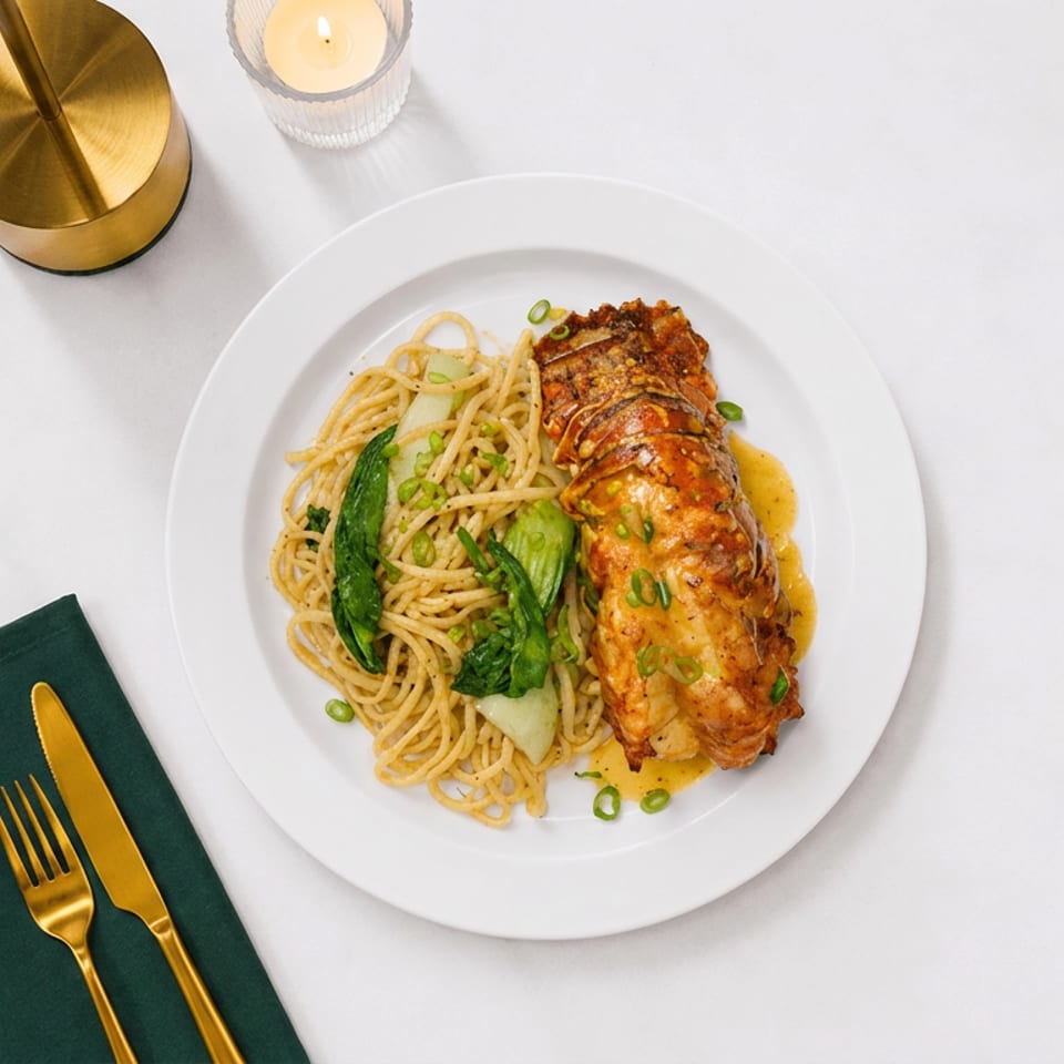 Overhead view of a plated lobster tail with noodles and bok choy, garnished with scallions, beside gold utensils and a candle.